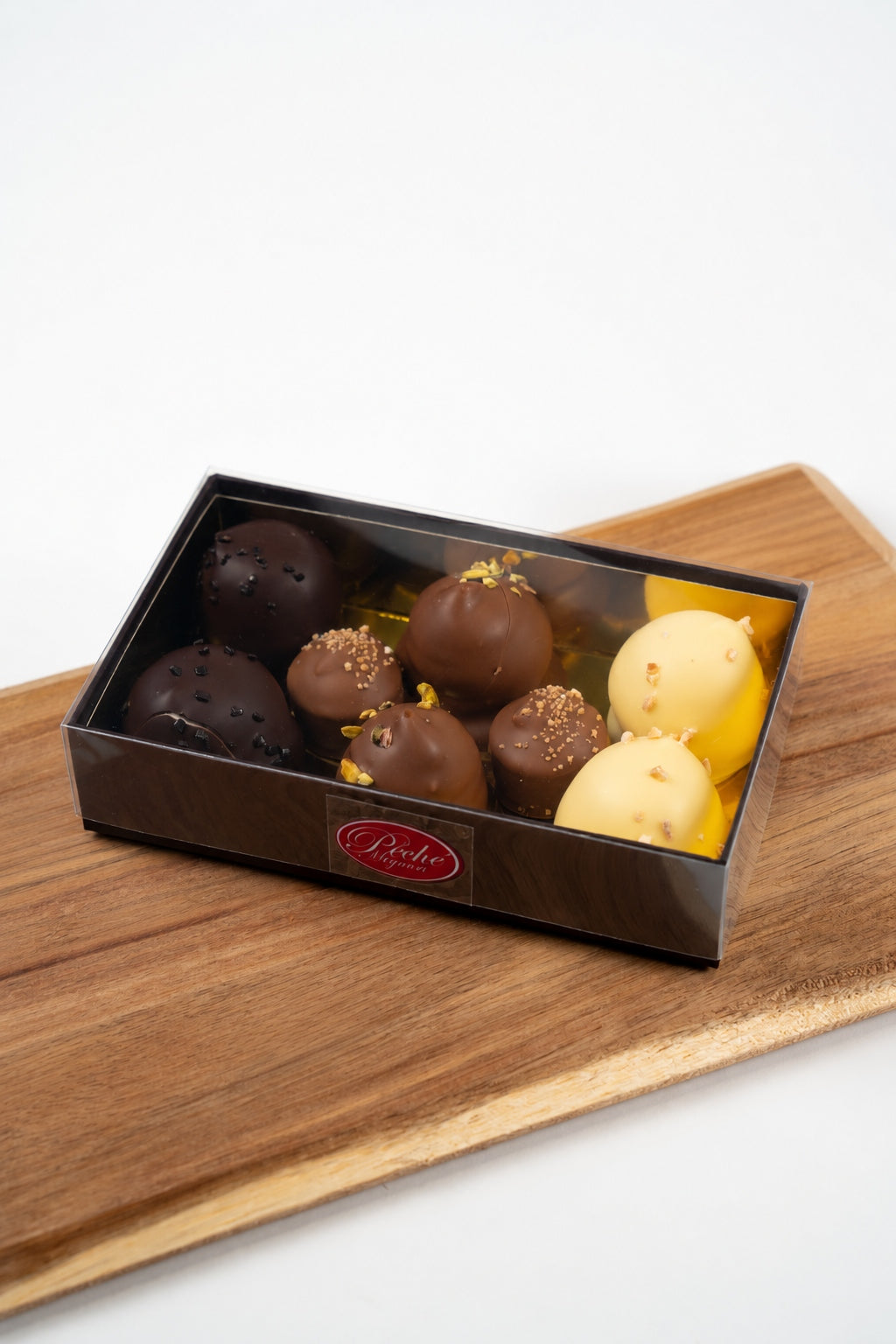Assortiment de bouchées en chocolat noir, au lait et blanc, décorées de graines et d’éclats, dans un coffret Péché Mignon à couvercle transparent posé sur une planche en bois.