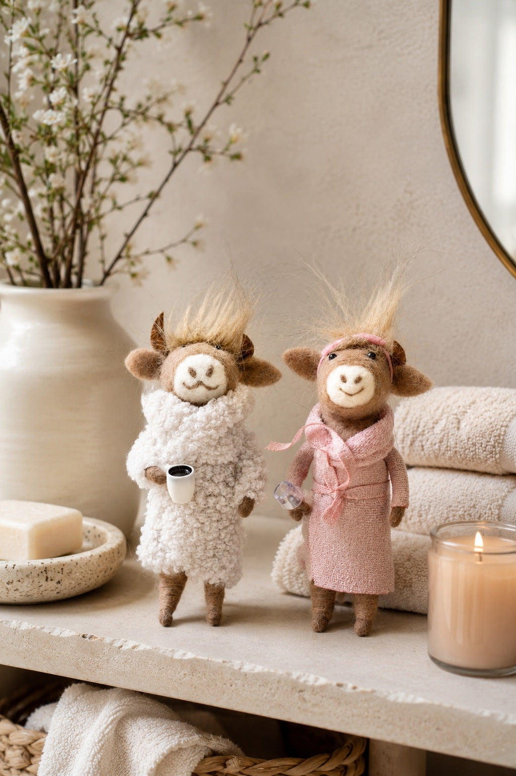 Deux figurines fantaisistes de vaches en feutrine, vêtues de peignoirs de spa, se tiennent sur une étagère de salle de bain à côté de serviettes pliées, d'une bougie allumée, de savon et d'un vase de fleurs blanches.