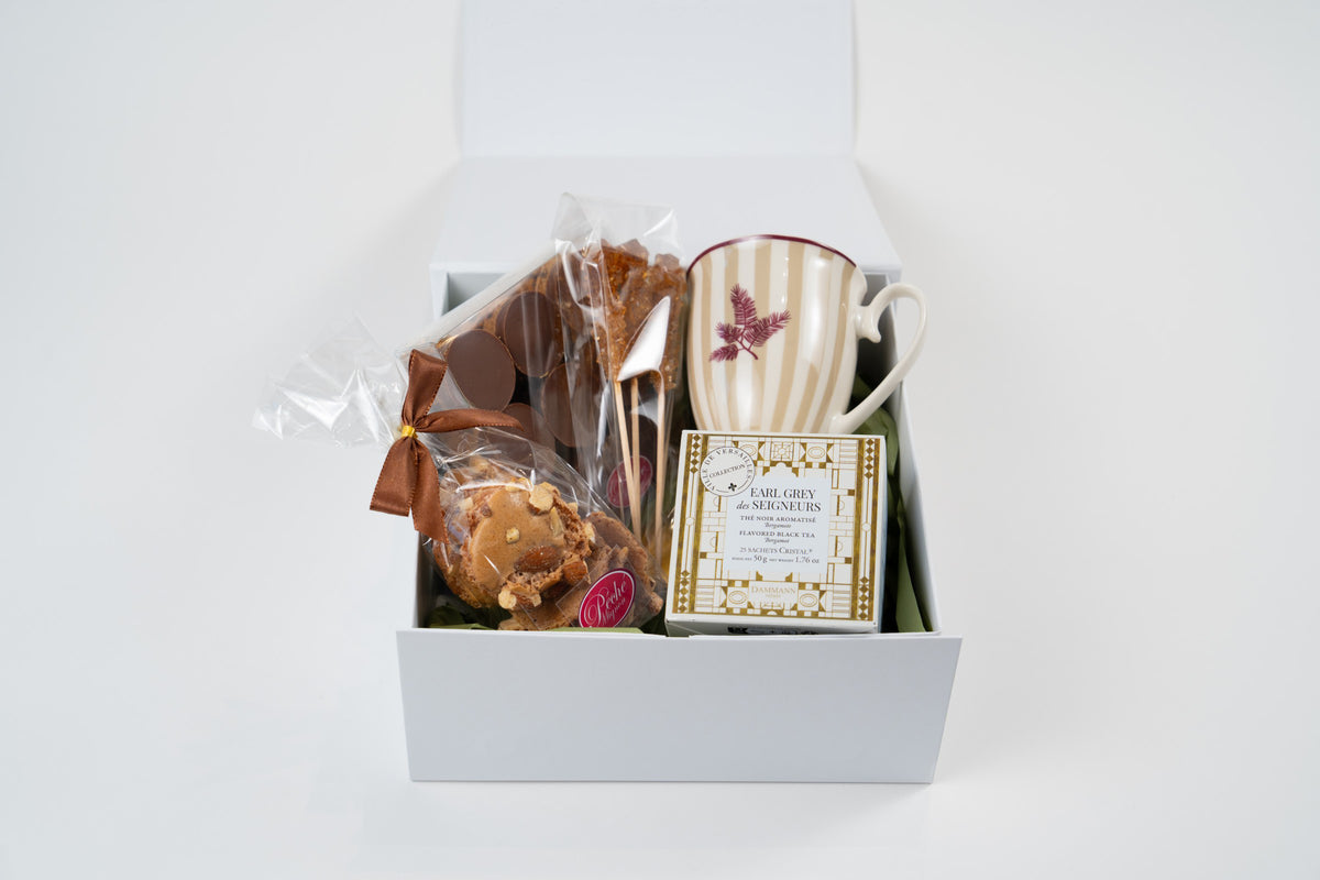 Open white tea-time gift box with almond meringues, chocolates, brown rock sugar sticks, Dammann Frères Earl Grey tea and a striped mug.