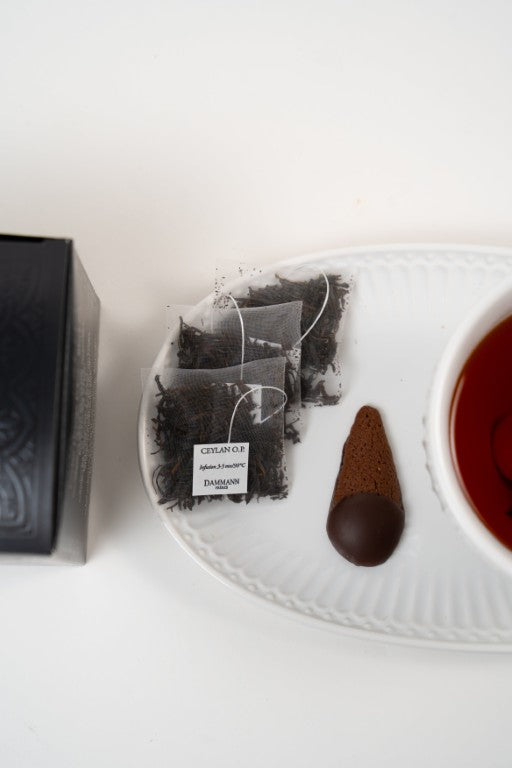 Dammann Frères Ceylan O.P. black tea sachets with cup of tea and chocolate biscuit on white plate.