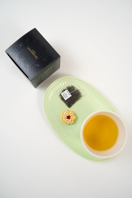 Cup of jasmine green tea with Dammann Frères tea box, tea sachet and biscuit on green plate.