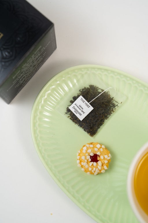 Dammann Frères jasmine green tea sachet with cup of green tea and biscuit on plate.