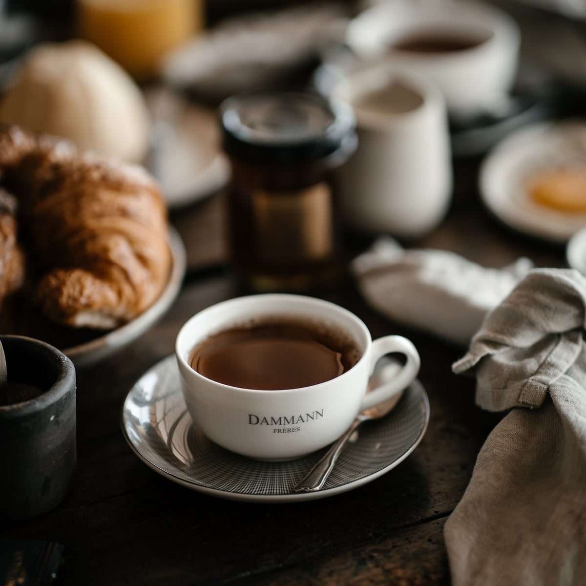 Elegant Dammann Frères tea cup on a rustic breakfast table with croissants and soft morning light in a luxury French café-style setting.