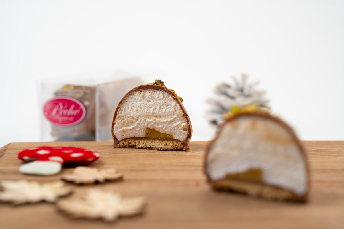Dubai-melocake met pistache en melkchocolade, doormidden gesneden zodat de marshmallow, pistachevulling en koekjesbodem zichtbaar zijn, met een tweede melocake op de voorgrond en een doos van Péché Mignon op de achtergrond.