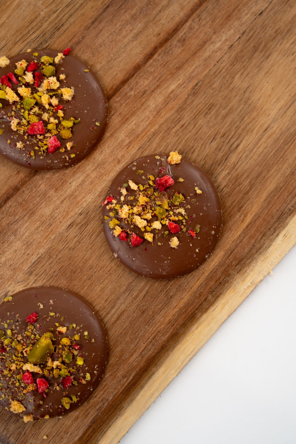 Belgian milk chocolate mendiant topped with pistachios and freeze-dried raspberry on a light background.