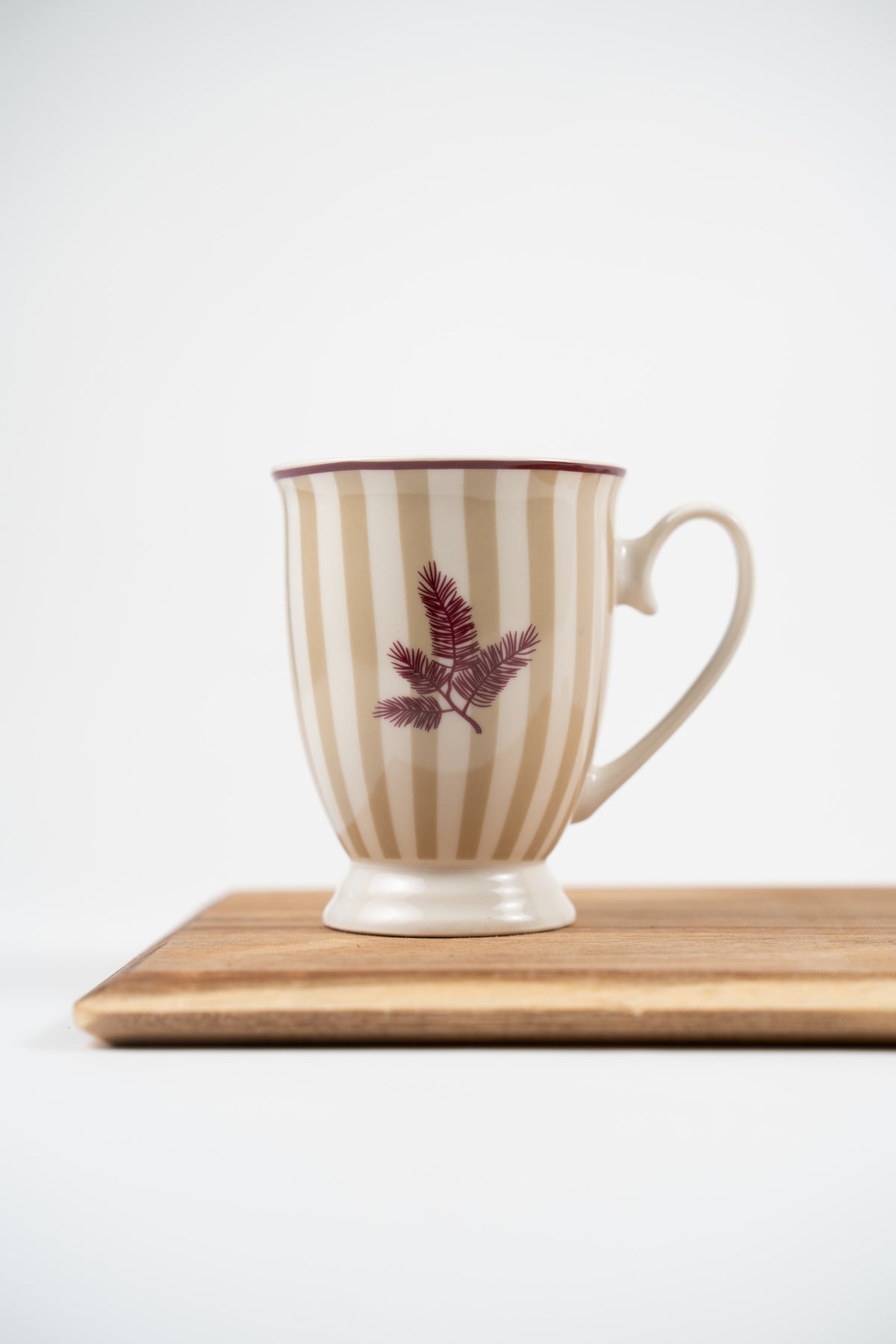 Naemi ceramic mug with beige and white stripes, a dark-red rim and delicate branch motif, on a wooden board.