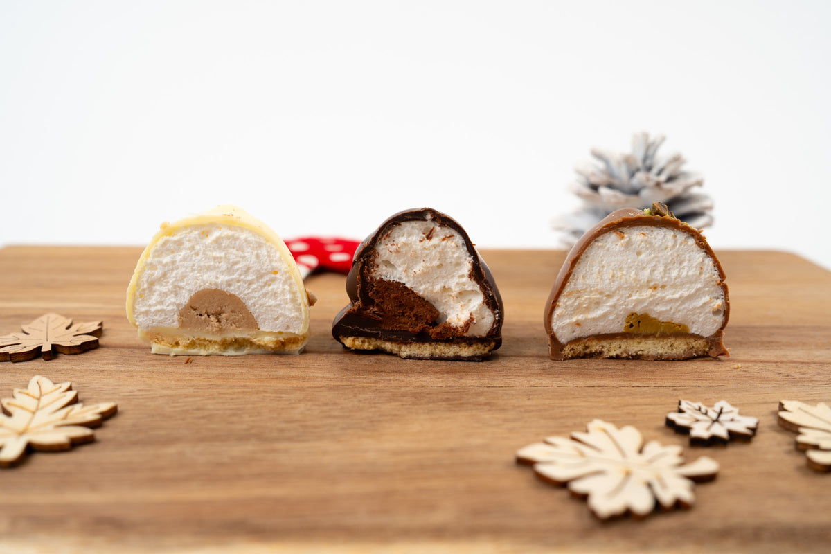 Trois Melocakes coupés en deux, de gauche à droite : chocolat blanc Kinder Bueno, chocolat noir mousse au chocolat et chocolat au lait Dubaï Pistache. Leurs intérieurs de guimauve et leurs bases biscuitées sont visibles sur une planche en bois.