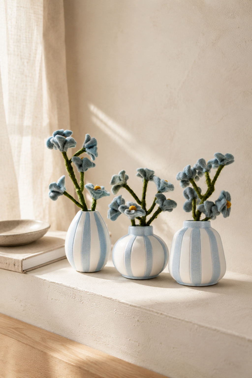 Three blue-and-white striped ceramic vases with handmade dusty blue felt flowers styled on a minimalist cream shelf with warm natural light.