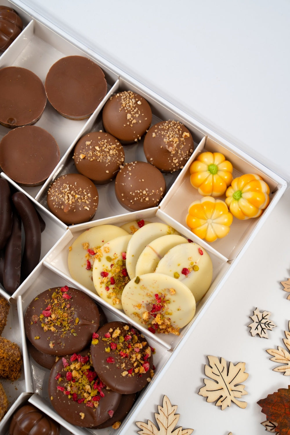 Close-up of an autumn gift box with round milk chocolate bites, white chocolate mendiants decorated with colourful pieces and yellow pumpkin-shaped chocolates.