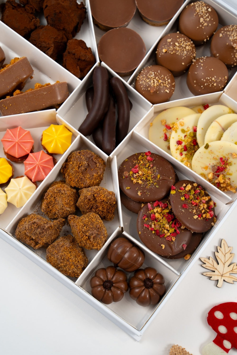 Close-up of a Péché Mignon chocolate assortment with coated truffles, milk chocolate bites, mendiants decorated with red and green pieces, and pumpkin-shaped chocolates.