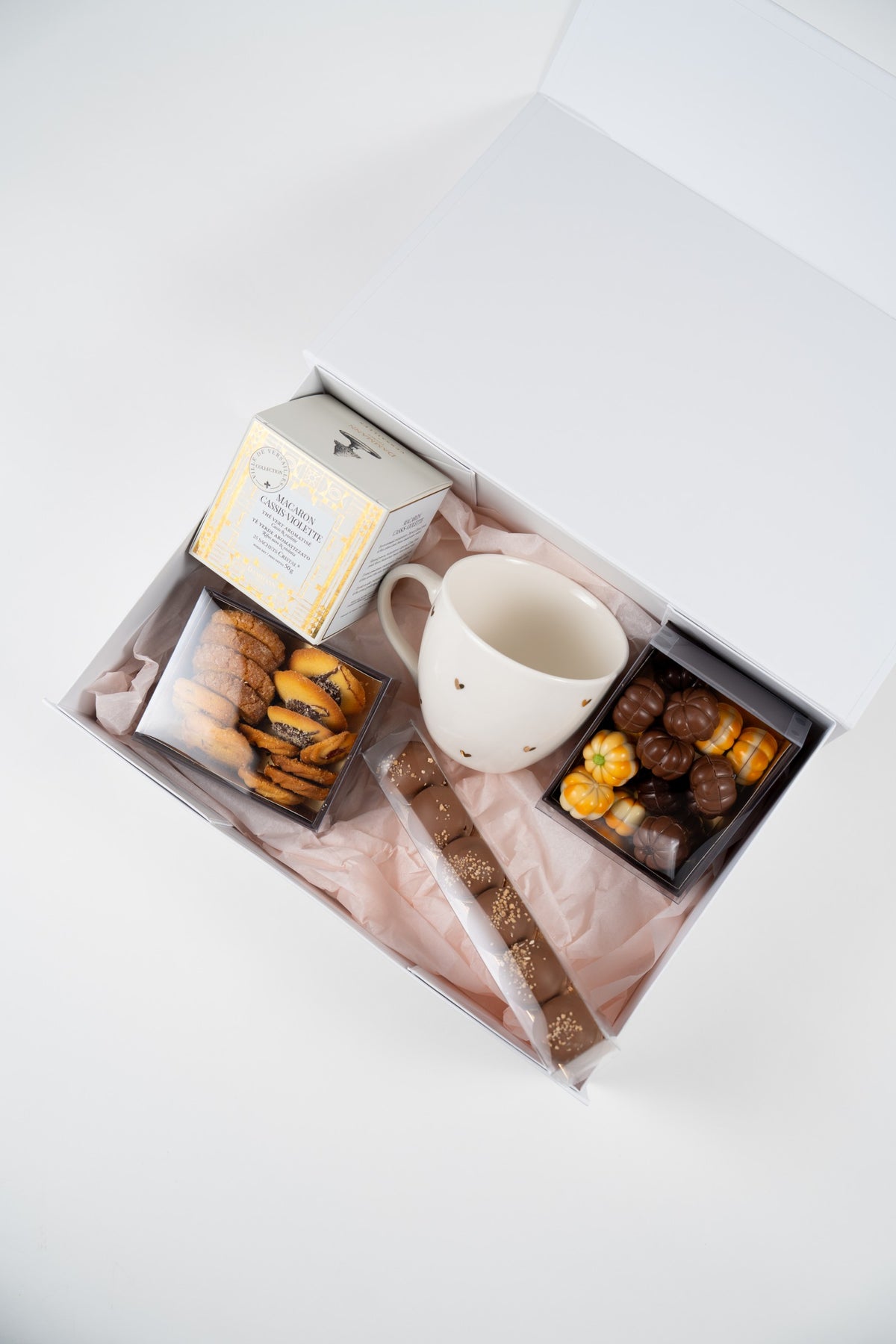Top view of a white autumn gift box with tea, mug, biscuits, pumpkin chocolates and a bar of milk-chocolate Mini Melocakes on pink tissue paper