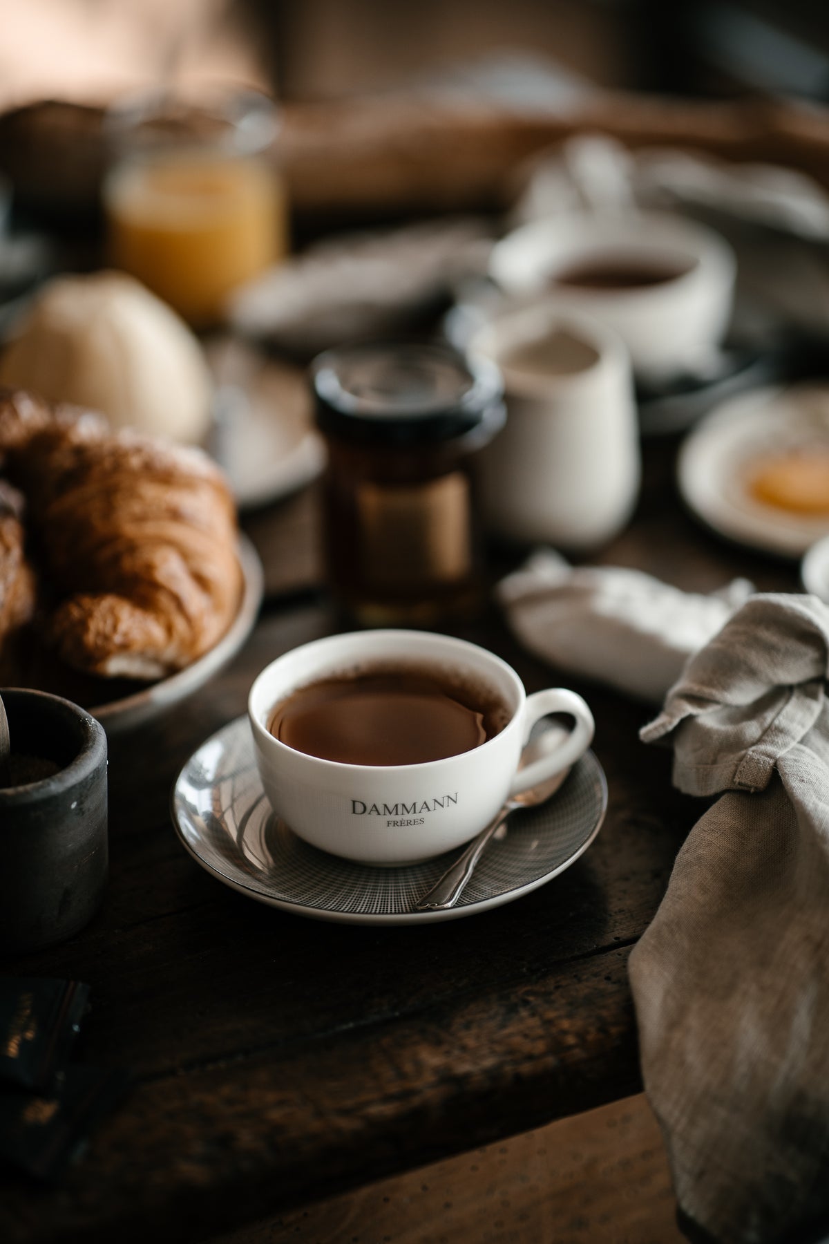 Elegant Dammann Frères tea cup on a rustic breakfast table with croissants and soft morning light in a luxury French café-style setting.