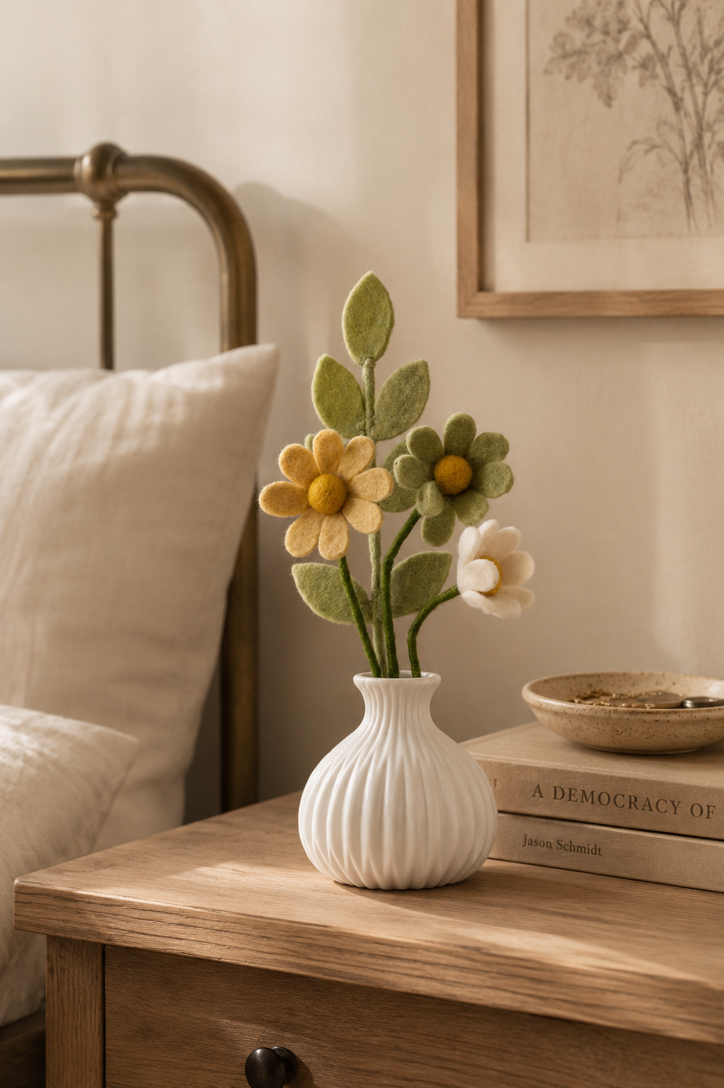 Small felt flower arrangement in a white ceramic vase styled on a wooden bedside table in a warm neutral bedroom interior.