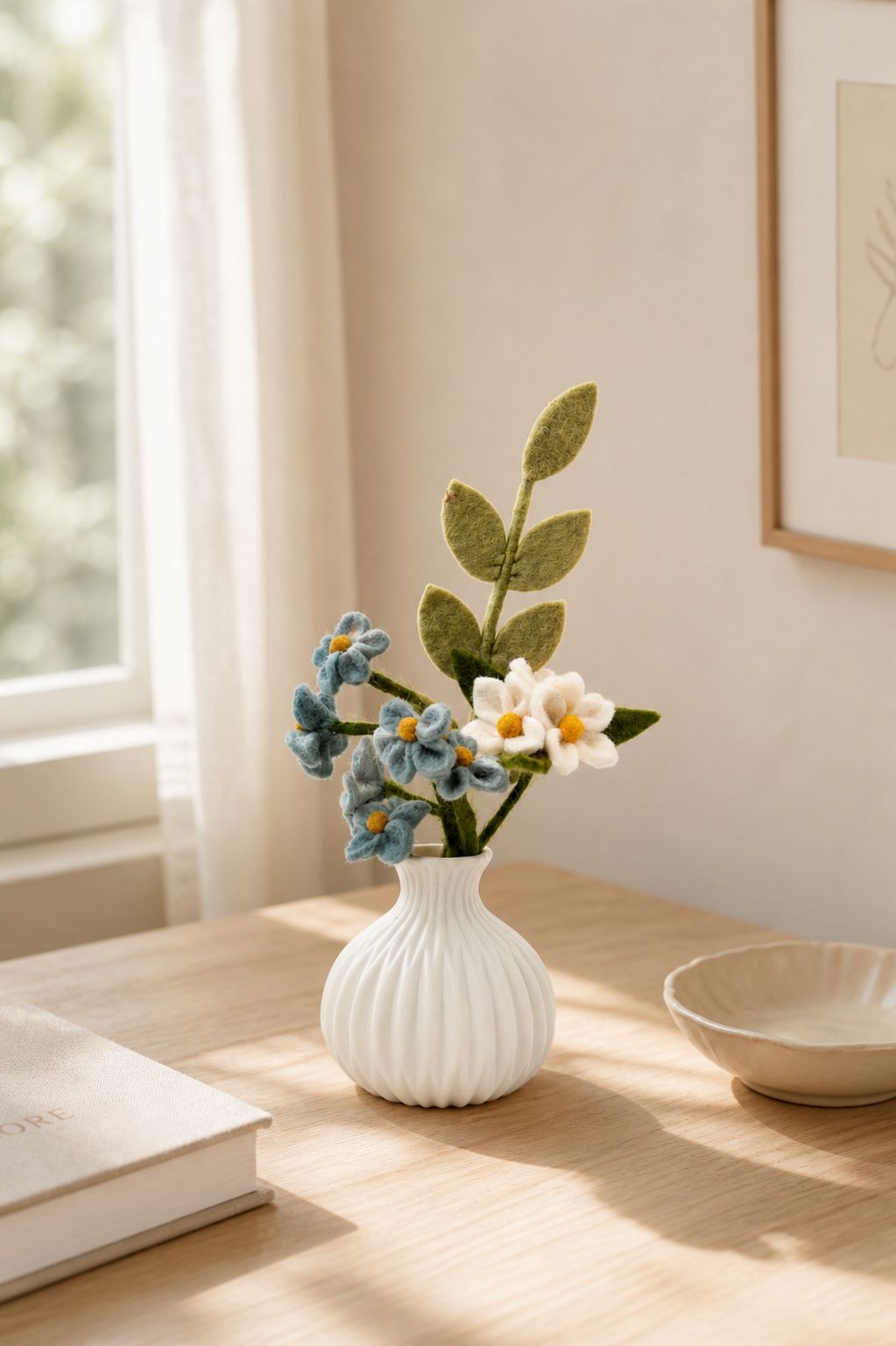 Handmade felt flower arrangement in a white ceramic vase styled on a light wood table in a warm minimalist Scandinavian-inspired interior.
