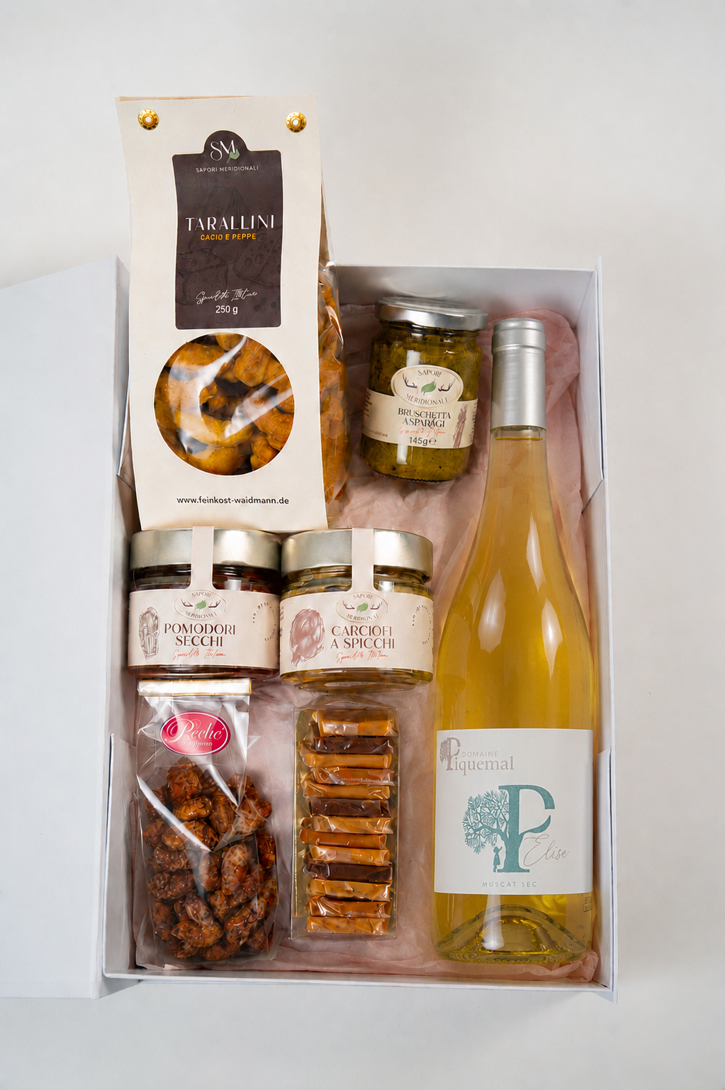 Luxury gourmet gift box featuring Italian delicacies including Tarallini crackers, asparagus bruschetta, preserved vegetables, caramel sweets, chocolate-coated nuts, and a bottle of Domaine Piquemal Elise Muscat wine arranged in a white box with pink tissue paper.