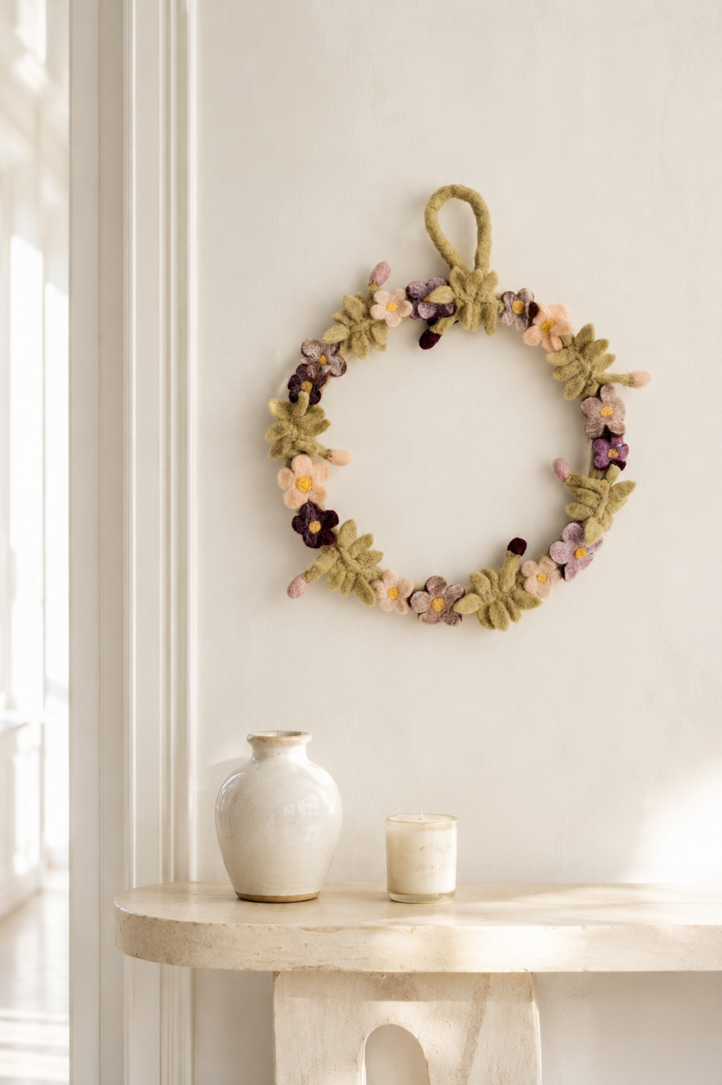 Handcrafted felt floral wreath hanging on an ivory wall above a minimal console with a ceramic vase and candle in a bright, elegant interior.