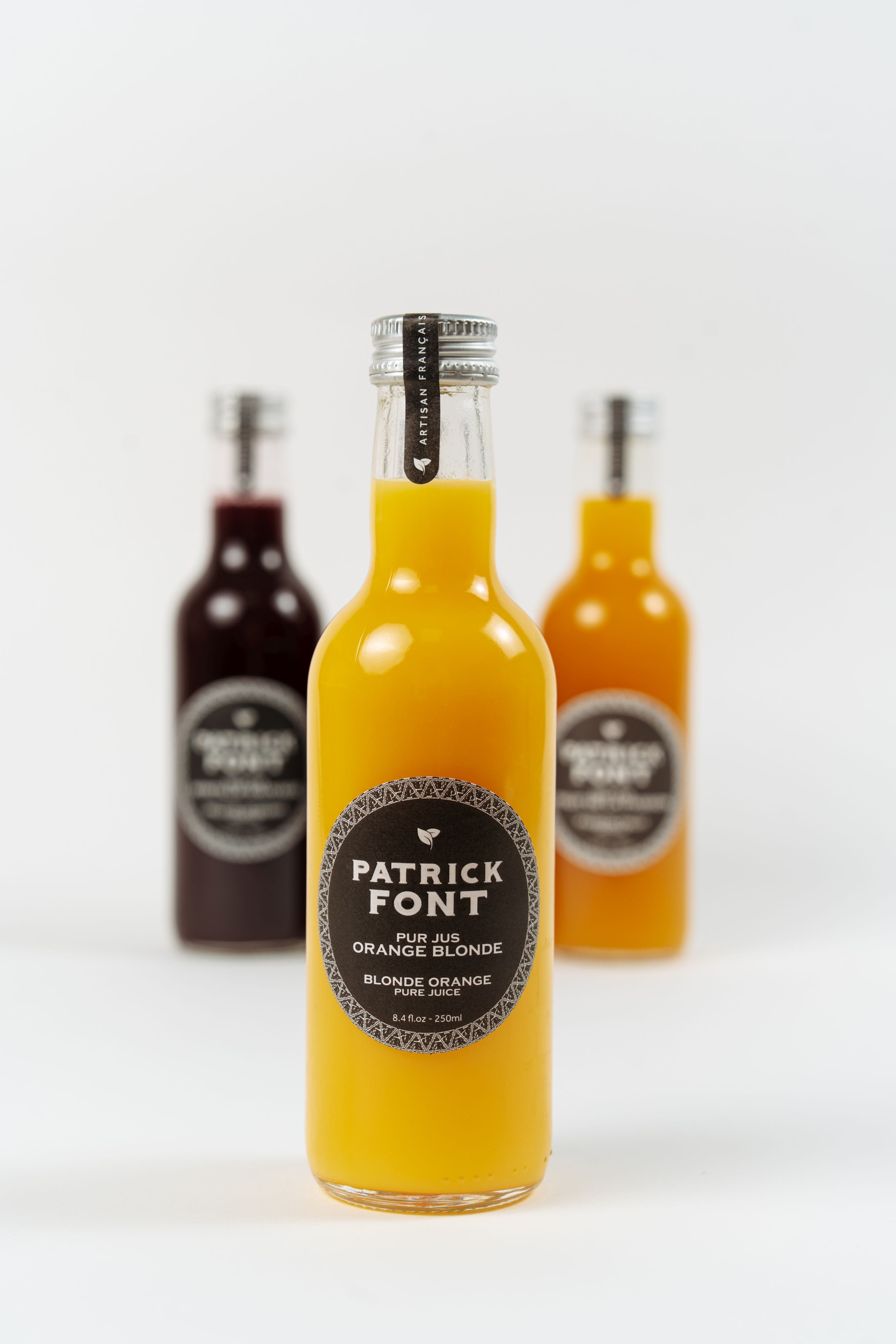 Bouteille de pur jus d’orange blonde Patrick Font avec bouteilles de jus en arrière-plan.