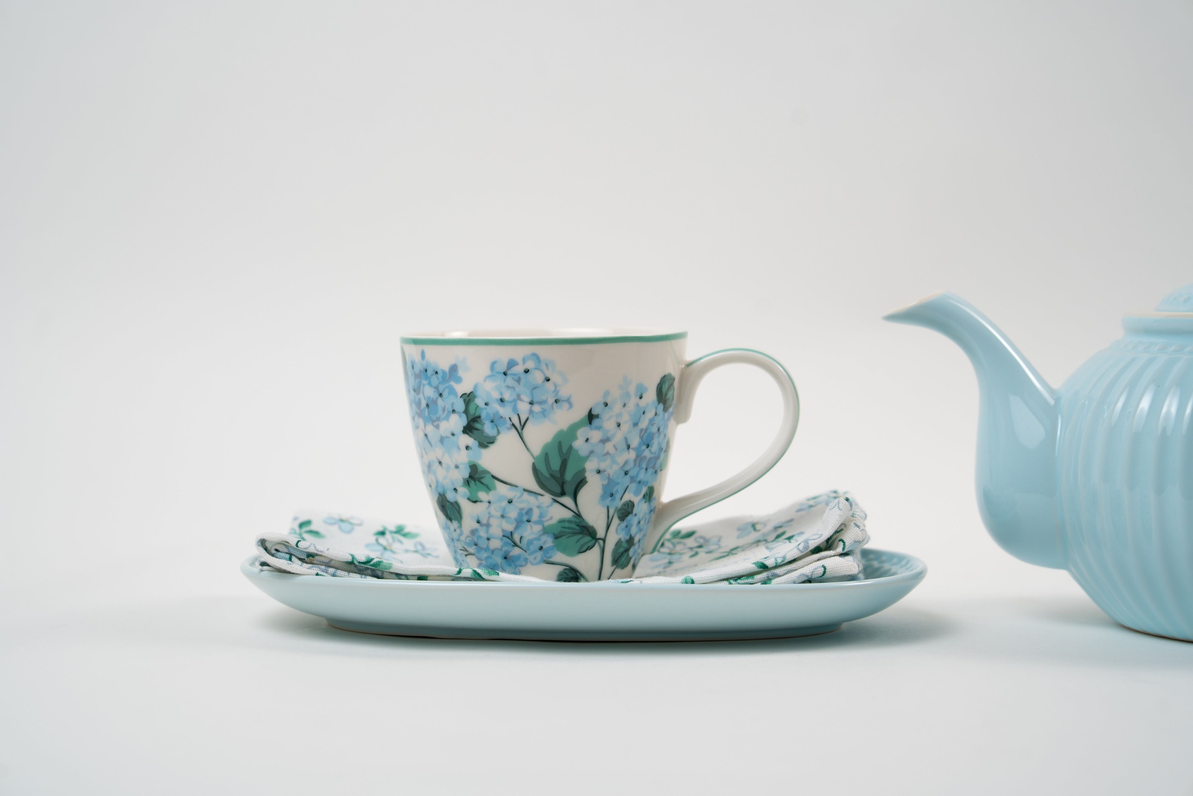 Tasse en porcelaine ornée de motifs floraux bleus, présentée avec sa soucoupe et sa serviette assortie. Un ensemble délicat pour une ambiance élégante et poétique.